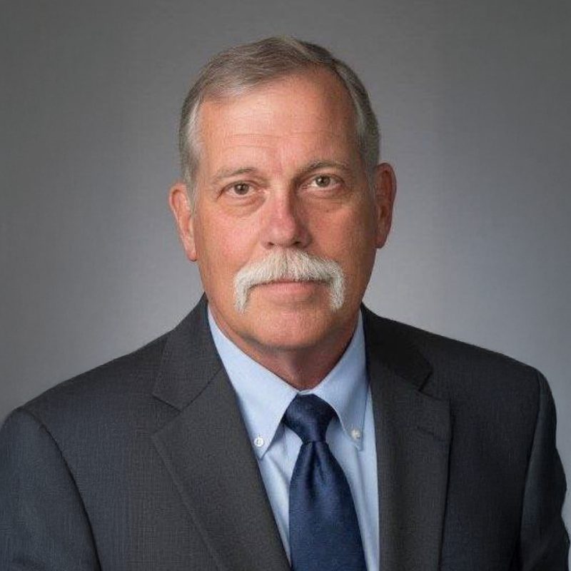 Steve Foley, in a charcoal suit and navy tie, with a white moustache, against a grey studio background